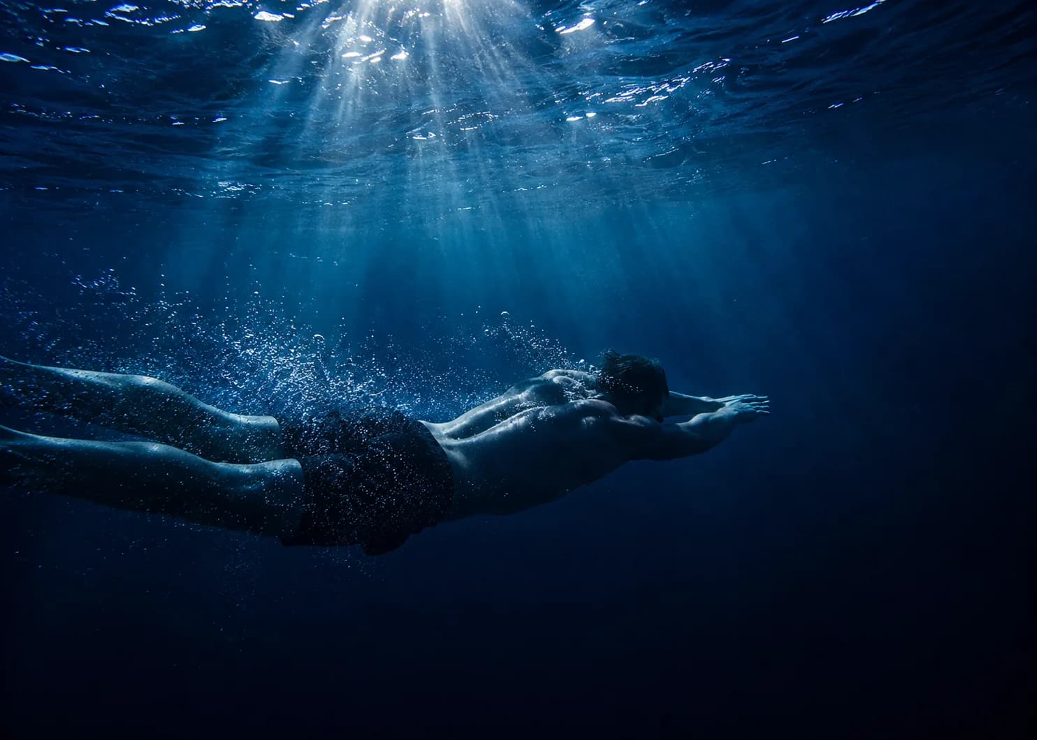 An underwater photograph of a swimmer gliding forward through deep blue water, sunlight beams filtering down from the surface